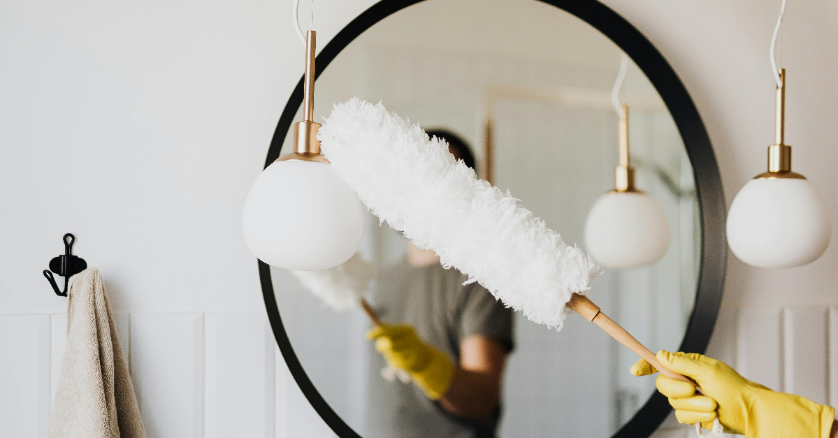 A person wearing yellow rubber gloves dusts a round mirror with a white duster in a bathroom.