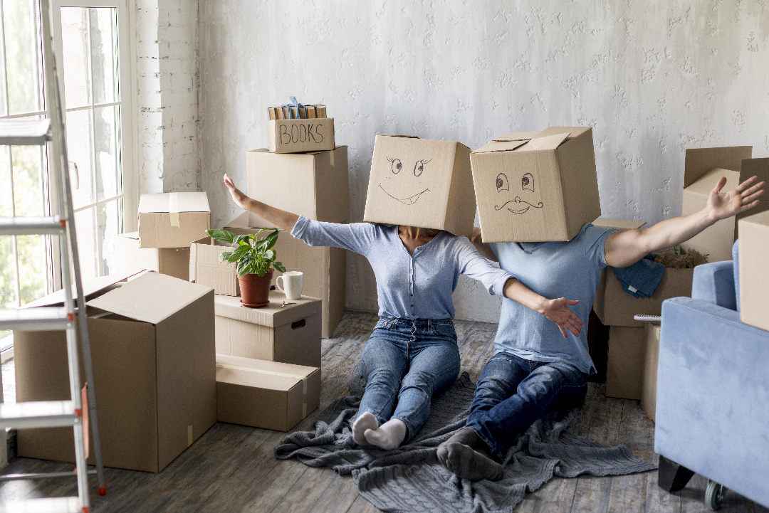 Two people sit on a floor surrounded by moving boxes, each with a box on their head featuring a drawn face. They are spreading their arms wide, and a potted plant rests nearby.