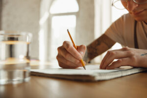 Person writing with a pencil on a notepad at a wooden table, with a glass of water nearby.