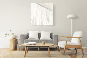 Minimalist living room with a gray sofa, two white cushions, a light wood chair, and a round coffee table. Floor lamp and small potted plant are near a large monochrome leaf wall art.