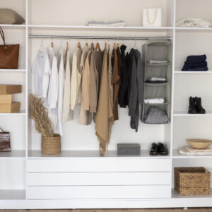 A neatly organized closet with white shelves, neutral-toned clothes hanging, folded garments, boxes, shoes, and baskets.